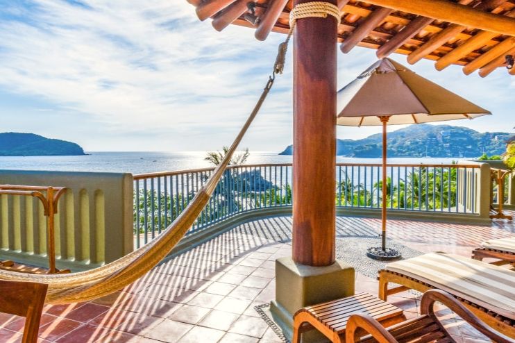 A serene ocean view from a sunny balcony featuring a hammock, wooden loungers, and a shaded table umbrella, surrounded by lush greenery and coastal hills, featured in the Hilton Grand Vacations Jackpot Sweepstakes