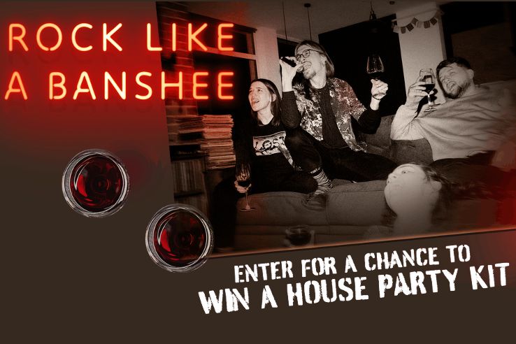 Three friends enjoy a lively evening at home, laughing and holding drinks. A bright neon sign above them reads "ROCK LIKE A BANSHEE," adding to the festive atmosphere, featured in the Banshee House Party Sweepstakes