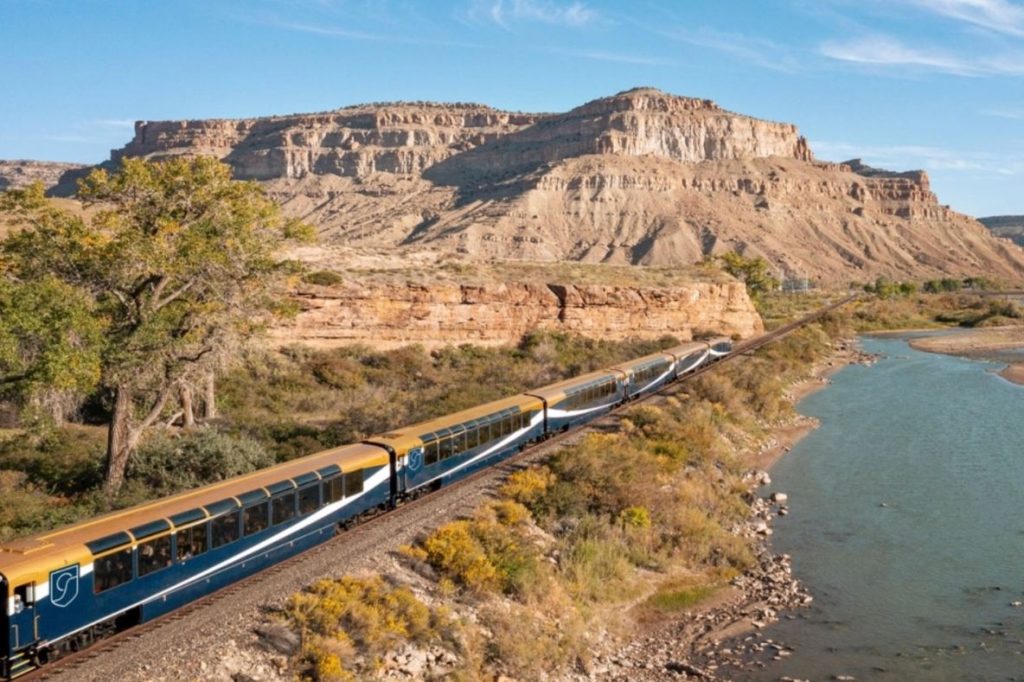 A colorful train winds along a rocky landscape next to a river, with towering cliffs and sparse vegetation under a clear blue sky, featured in the Canyon Spirit 3-Day Rail Sweepstakes