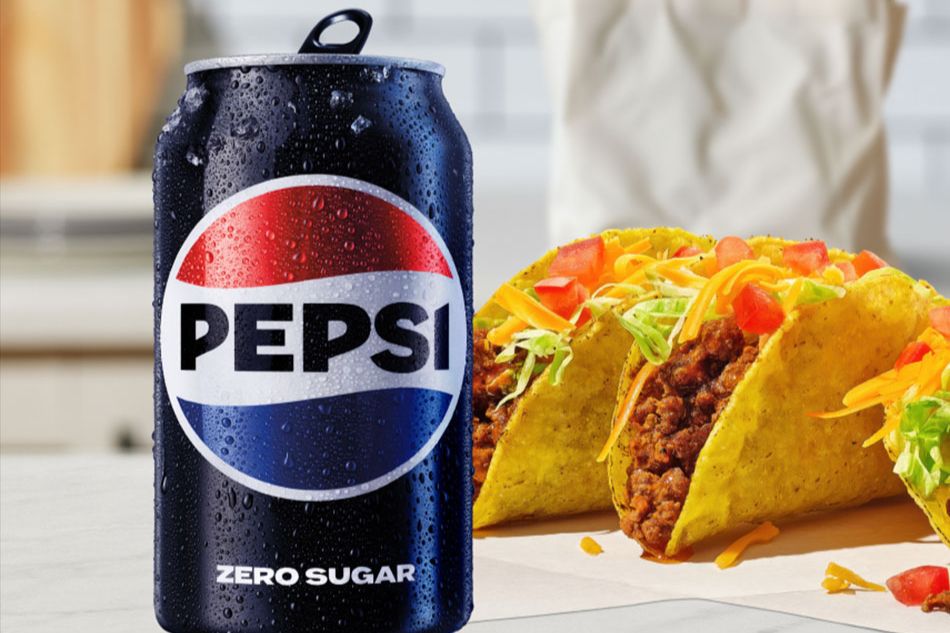 A chilled can of soda sits next to two loaded tacos, showcasing vibrant ingredients like cheese, lettuce, and tomatoes against a soft kitchen backdrop, featured in the Pepsi Groceries for a Year Sweepstakes