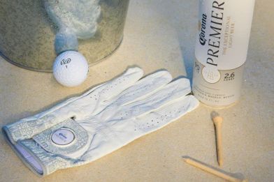 A white golf glove lies on a sandy surface beside a golf ball, a golf tee, and a can of premium beer, suggesting a relaxed day on the golf course, featured in the Corona Golf 2026 Sweepstakes