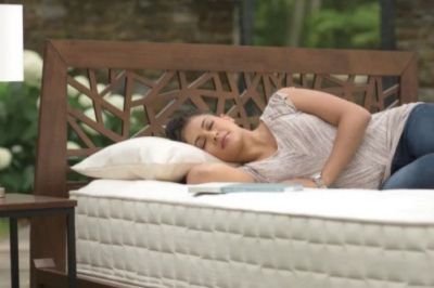 A woman peacefully sleeps on a stylish, patterned bed frame with a plush mattress, surrounded by a serene outdoor setting filled with greenery, featured in the TheRoundup Naturepedic Mattress Sweepstakes