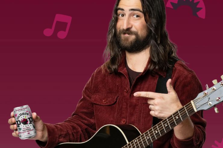 A man with long hair and a beard holds a soda can while playfully pointing with his other hand. He stands with an acoustic guitar against a colorful, patterned background, featured in the Culture Pop Noah Kahan Tickets Sweepstakes
