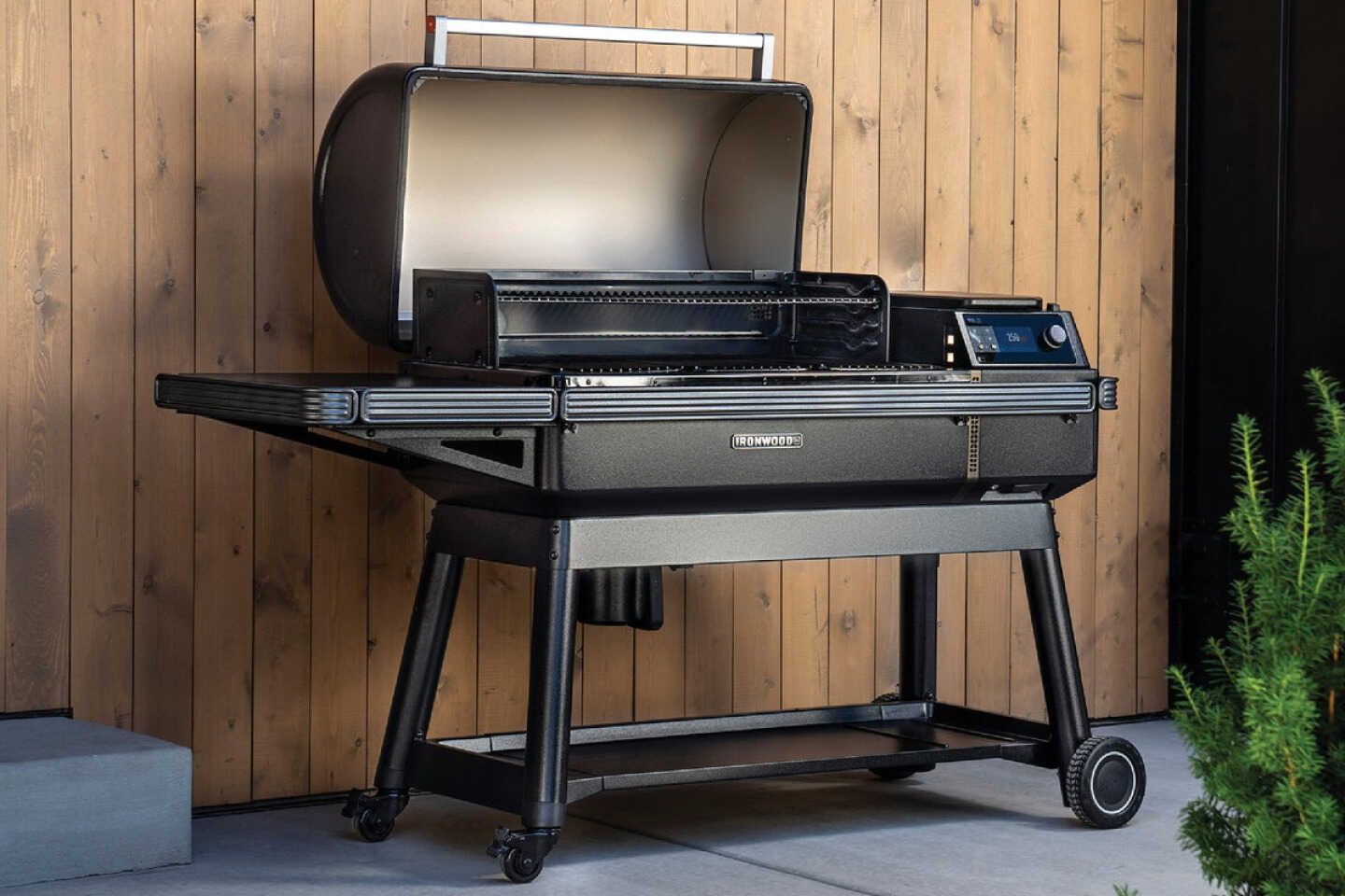 A sleek black grill with an open lid stands on a patio, set against a wooden wall. It features a spacious cooking area and wheels for mobility, surrounded by greenery, featured in the Michael David Winery Traeger Grill Sweepstakes