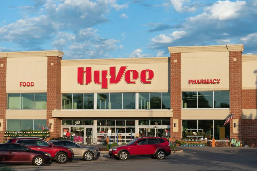 A bustling grocery store facade features large windows, signs for "Food" and "Pharmacy," and parked cars in front. The sky showcases scattered clouds, creating a vibrant atmosphere, featured in the Hy-Vee Easter Meal Pack Sweepstakes