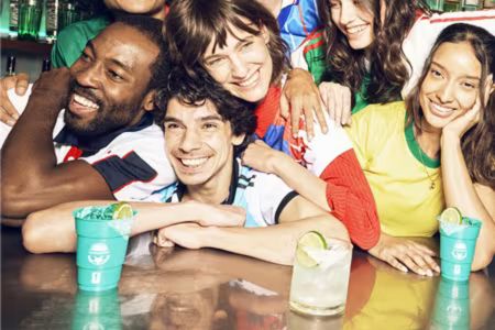 A diverse group of friends smiles and poses together at a bar, wearing colorful jerseys. Cocktails are placed on the counter in front of them, capturing a lively, celebratory atmosphere, featured in the Casamigos FIFA World Cup Sweepstakes