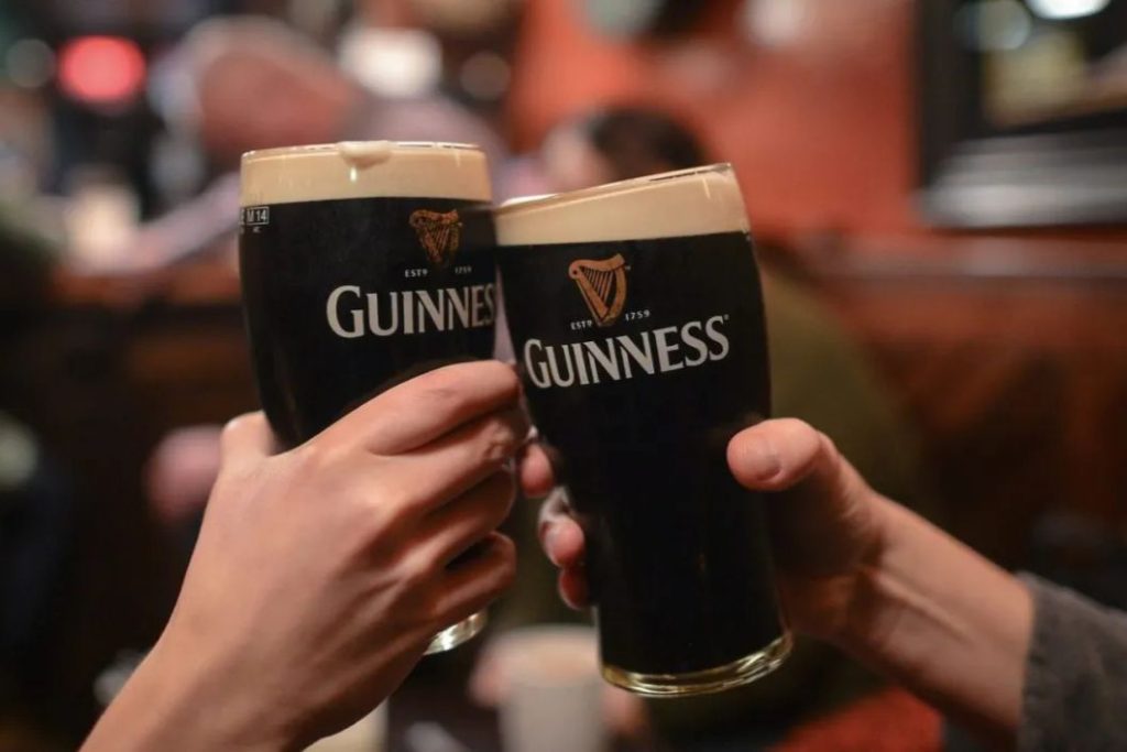 Two hands toast with pints of dark beer in a lively pub setting, highlighting a moment of celebration and camaraderie among friends, featured in the Guinness St. Patrick's Day Countdown Sweepstakes