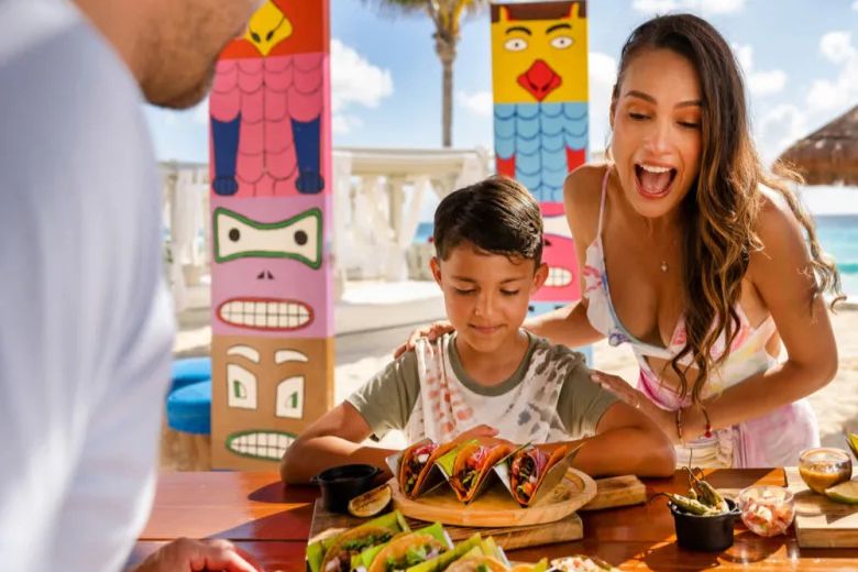 A woman joyfully interacts with a boy at a vibrant beachside table, showcasing a delicious spread of tacos and sides, surrounded by colorful totem-like decorations, featured in the Sunscape Cancun Fun in the Sun Sweepstakes