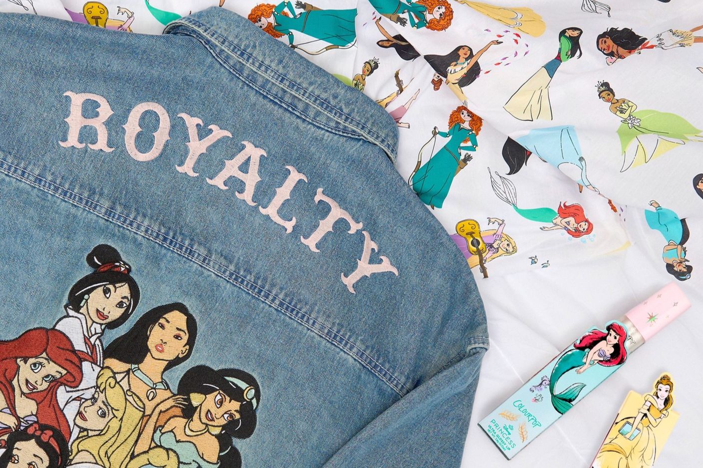 A denim jacket adorned with illustrations of various princesses and the word "ROYALTY" on the back is laid out with a patterned fabric and a beauty product nearby, featured in the Colourpop Ultra Glossy Lip Sweepstakes