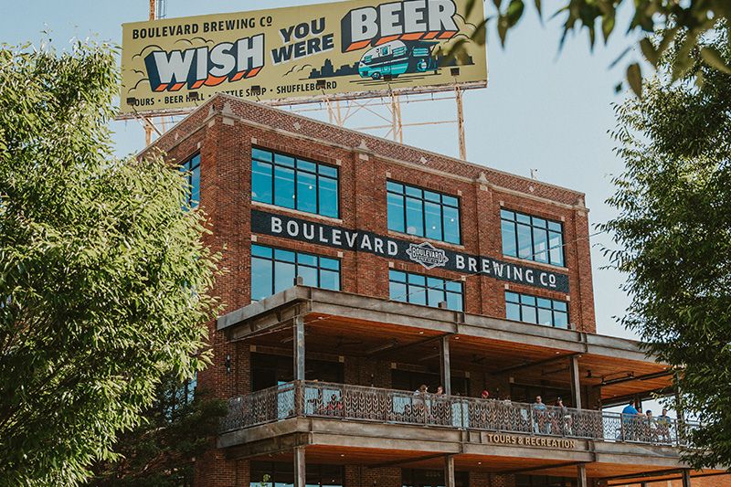 A brick building with large windows features guests enjoying a balcony. Above, a billboard promotes Boulevard Brewing Co. in a sunny, tree-lined setting, featured in the Boulevard Royals Fly Away Sweepstakes