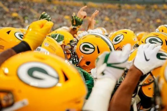 A close-up of a huddle of football players wearing yellow helmets with a green logo, engaged in a pre-game chant, with fans visible in the blurred background, featured in the American Family Insurance Packers Draft Sweepstakes