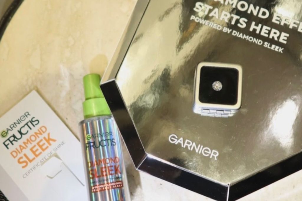 A sleek, reflective display features a diamond-sparkling compact and a colorful spray bottle, both promoting a hair product line, resting on a smooth surface, featured in the Garnier Diamond Sleek Social Sweepstakes
