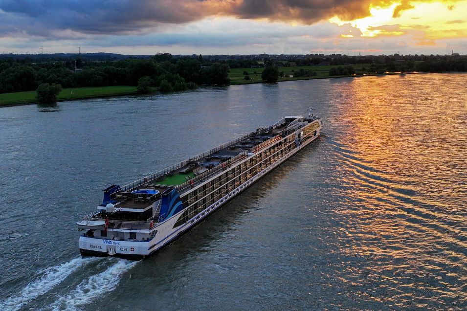 A large river cruise ship sails smoothly on a tranquil waterway at sunset, casting reflections of golden hues on the water, with lush green banks in the background, featured in the VIVA Cruises Set Sail and Let's Go Sweepstakes