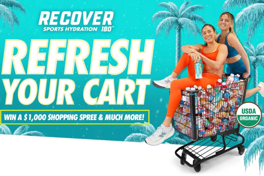 Two women are playfully posing on a shopping cart filled with colorful sports drinks, set against a tropical background with palm trees. They exude energy and fun, featured in the RECOVER 180 Gift Card Sweepstakes