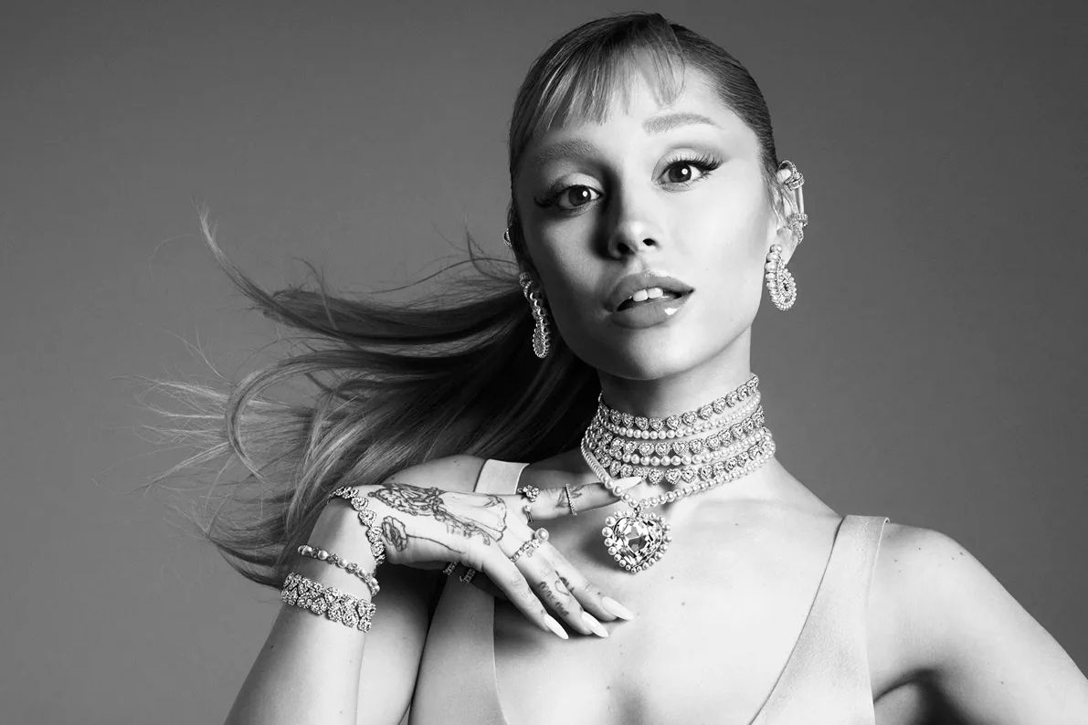 A glamorous individual poses elegantly with flowing hair, adorned in sparkling jewelry including earrings, a choker, and bracelets, exuding confidence and style in a monochrome setting, featured in the Swarovski Ariana Grande Sweepstakes