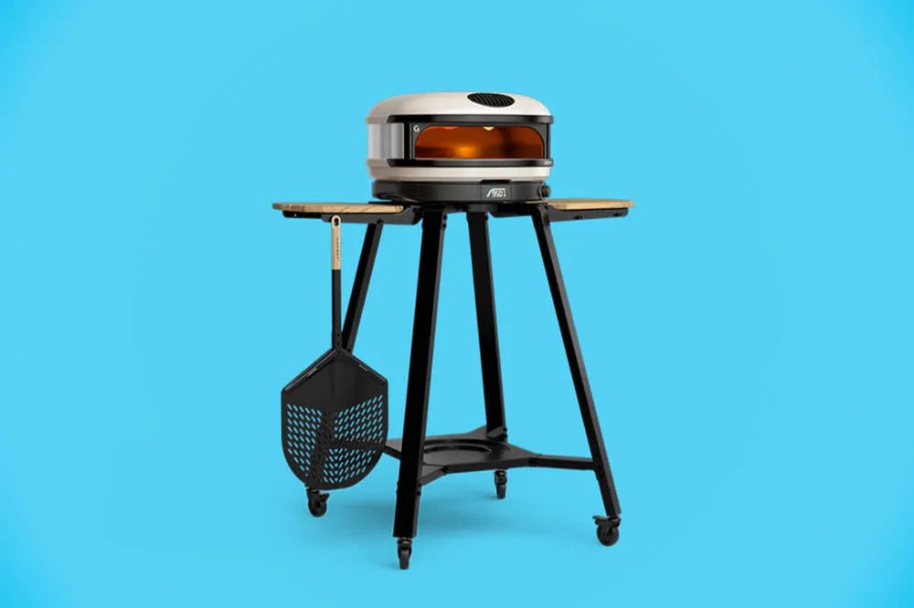 A compact outdoor pizza oven on a black cart with wooden shelves, featuring a metal hood and clear viewing window. A pizza peel hangs beside the oven for easy access. The background is light blue, featured in the The Strategist Gozney Pizza Oven Sweepstakes