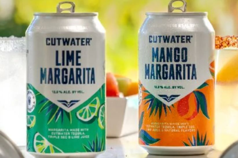 Two cans of margarita drinks sit side by side on a sunlit table. One can features a vibrant lime design, while the other showcases a colorful mango theme, with fresh fruit in the background, featured in the Cutwater $5,000 Patio Makeover Sweepstakes