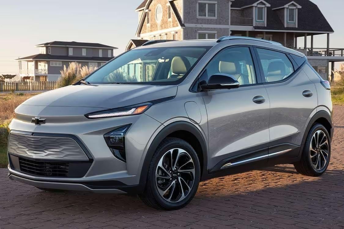 A sleek silver SUV is parked on a brick path, with a coastal house visible in the background. The vehicle has modern lines and stylish wheels, reflecting a contemporary design, featured in the Schilling Cider Chevy Bolt Sweepstakes