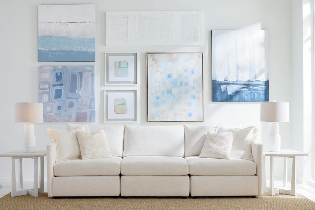 A cozy white sofa with decorative pillows sits in a bright room, adorned with various abstract art pieces on the wall and paired with stylish side tables and lamps, featured in the Bassett 15000 Sweepstakes