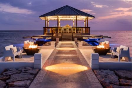 A serene outdoor venue features a wooden pavilion on a pier, surrounded by glowing fire pits and inviting lounge chairs, all set against a tranquil ocean at sunset, featured in the Sandals and Beaches Giveaway Q2, 2026 Sweepstakes