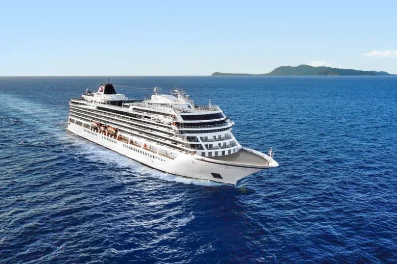 A large cruise ship sails smoothly through a vibrant blue ocean, leaving a wake behind it. In the distance, a lush green island is visible under a clear sky, featured in the Viking 8-Day Journey Sweepstakes