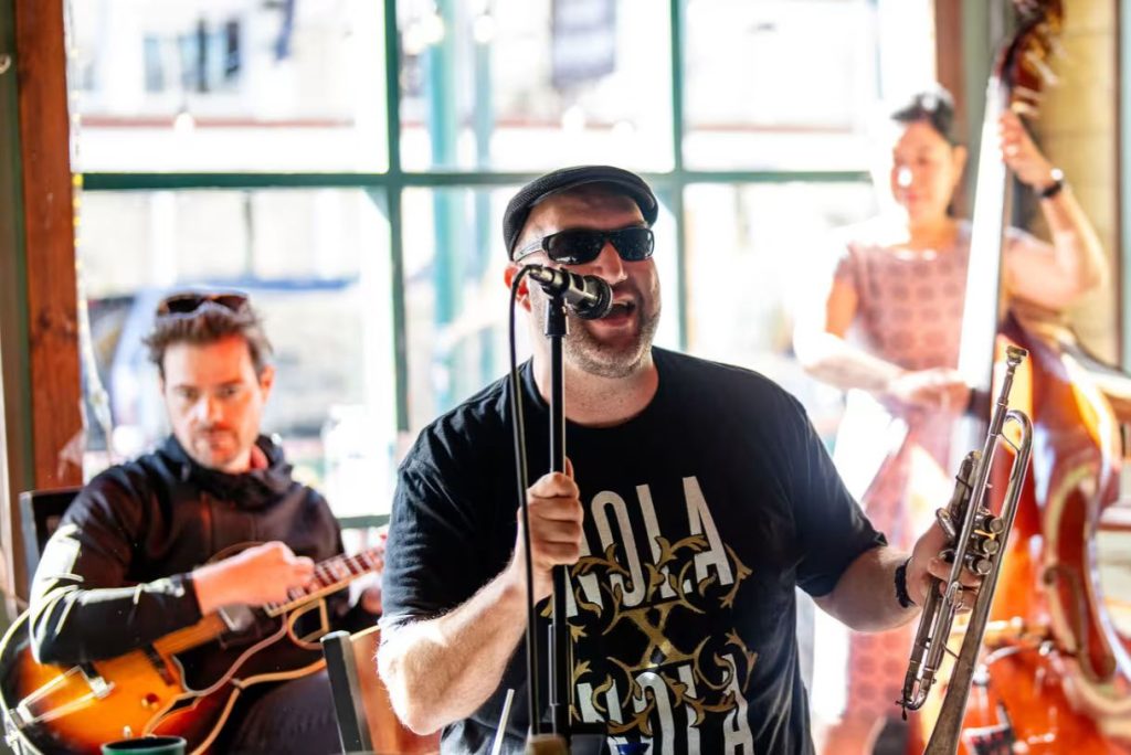 A male musician passionately sings into a microphone while holding a trumpet, accompanied by a guitarist and a woman playing a double bass in a lively indoor setting, featured in the NOLAxNOLA 2026 Sweepstakes