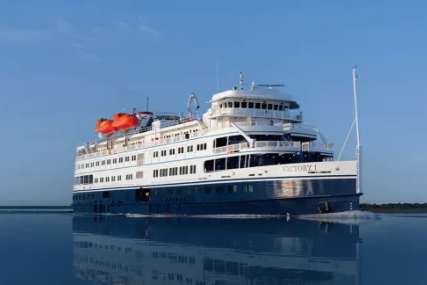 A large cruise ship named Victory I navigates through calm waters, with vibrant flags flying from its upper deck against a clear blue sky, reflecting on the water's surface, featured in the Victory Cruise Lines Discover the Great Lakes Sweepstakes