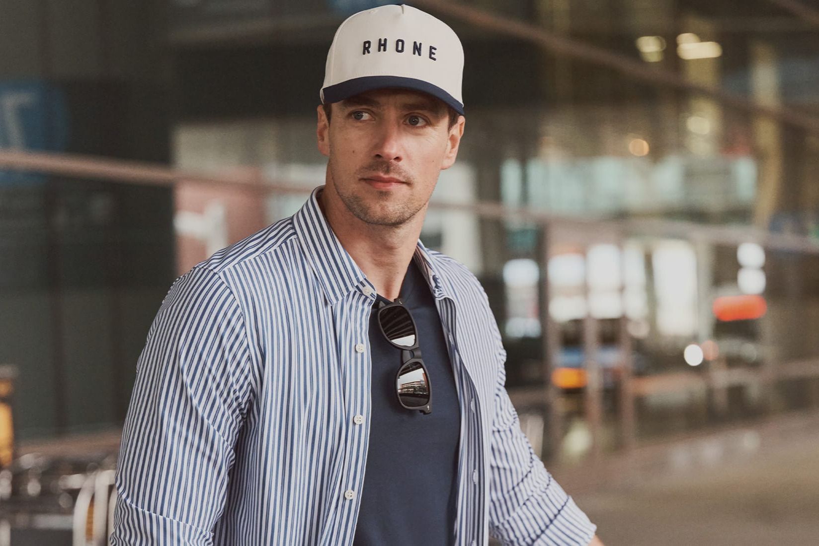 A casually dressed man walks through a modern airport terminal, wearing a striped shirt and a cap. He has sunglasses tucked into his shirt while glancing off-camera, featured in the Rhone Commuter Upgrade Sweepstakes