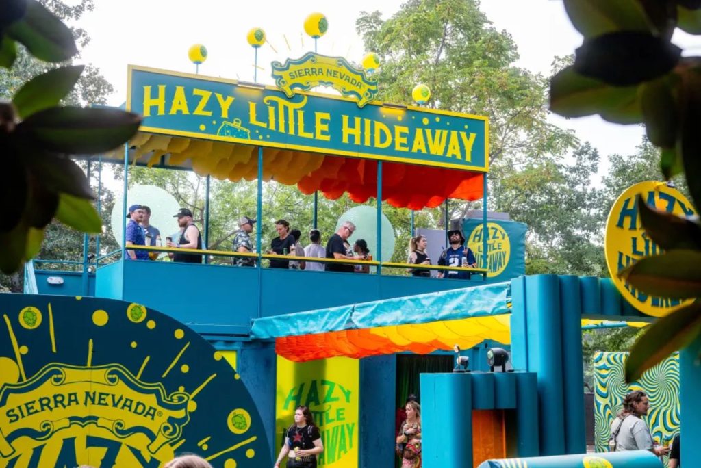 A vibrant outdoor venue features a colorful deck labeled "Hazy Little Hideaway," where people mingle and enjoy drinks amidst trees and bright decor, featured in the Sierra Nevada EDC Ticket Sweepstakes