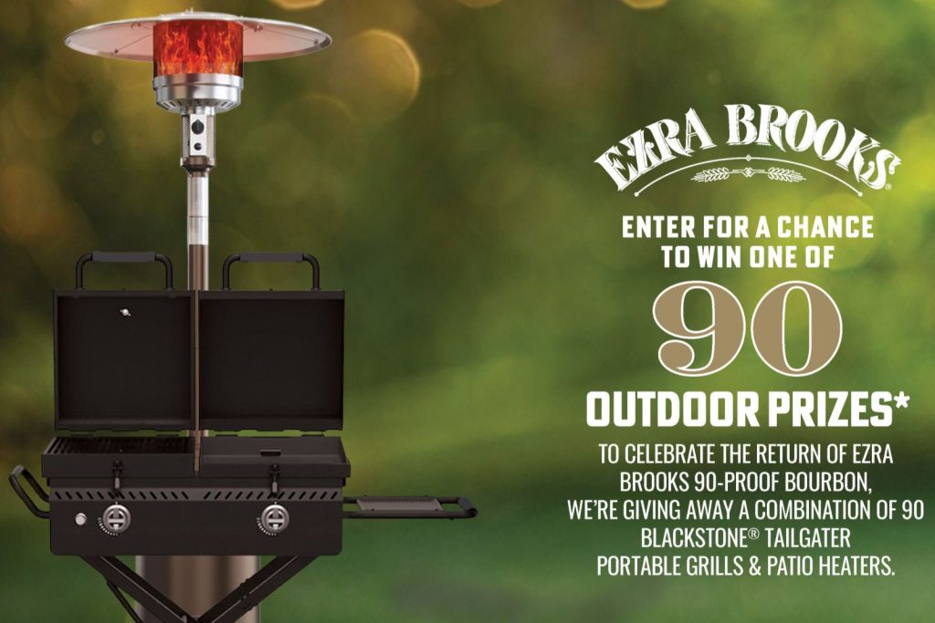 A shiny, silver outdoor patio heater stands beside a Blackstone tailgate grill, set against a blurred green background, creating an inviting atmosphere for outdoor gatherings, featured in the Ezra Brooks Grill & Heater Sweepstakes