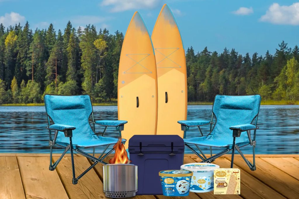 Two yellow surfboards stand upright by a serene lake, accompanied by blue camping chairs, a cooler, and a portable stove with a small flame, creating a cozy outdoor scene, featured in the Kemps Headed North Sweepstakes