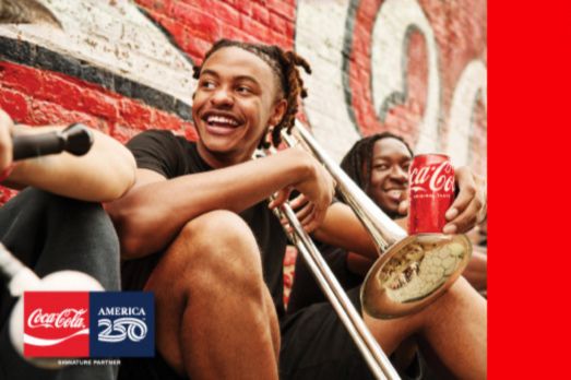 Two young men sit against a vibrant brick wall, one holding a trombone and the other enjoying a Coke, both smiling and engaging in a fun moment together, featured in the Coca-Cola Essence Festival Sweepstakes