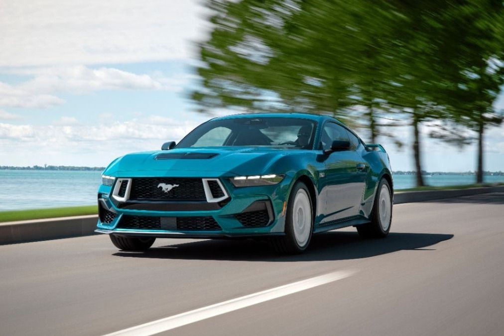 A sleek, turquoise sports car speeds along a scenic roadway beside a tranquil lake, with blurred trees in the background enhancing the sense of motion, featured in the Straight Talk Mustang Sweepstakes