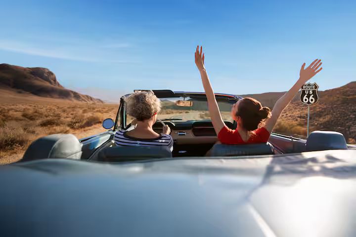 Two women enjoy a scenic drive in a convertible along a desert highway, with one sitting back and raising her arms in excitement, surrounded by mountains and blue skies, featured in the Wyndham Route 66 Roadtrip Sweepstakes