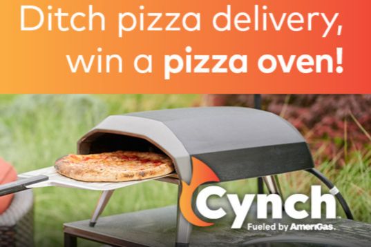 A pizza is being pulled from a sleek outdoor pizza oven, set against a vibrant garden backdrop, inviting viewers to embrace homemade pizza over delivery, featured in the Cynch Pizza Oven Sweepstakes