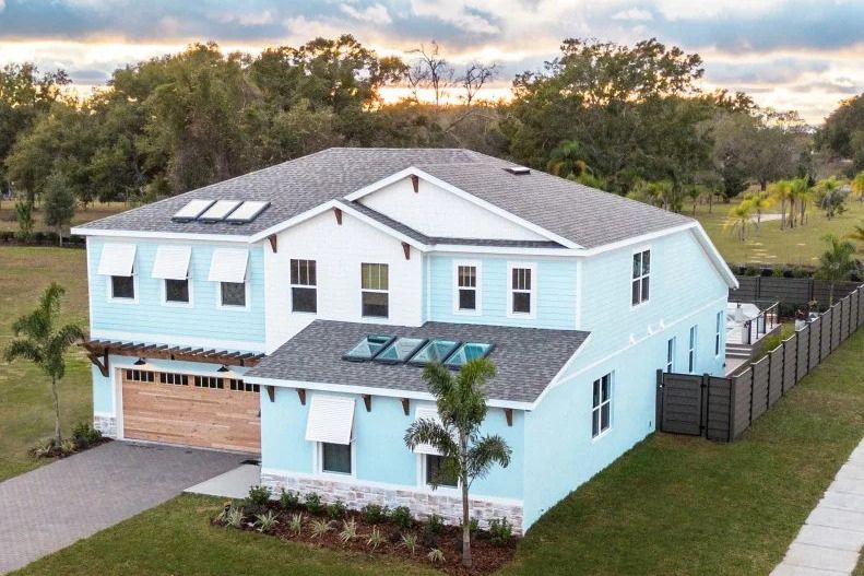 A large, light blue house with multiple windows and a wooden garage door, surrounded by lush greenery and a well-maintained yard under a cloudy sky, featured in the HGTV Smart Home 2026 Sweepstakes