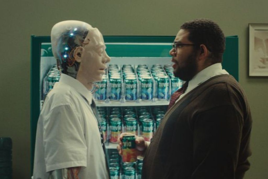 A humanoid robot and a man stand face to face in an office setting, with a vending machine filled with cans in the background. The man holds a can while they engage in conversation, featured in the Zevia Soda for a Year Sweepstakes