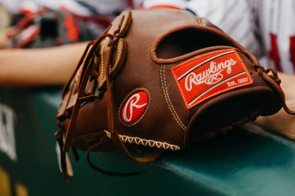 A close-up of a brown baseball glove resting on a green bench. The glove features red and white branding details, suggesting it's a high-quality Rawlings model, featured in the Sport Clips Playmaker Giveaway