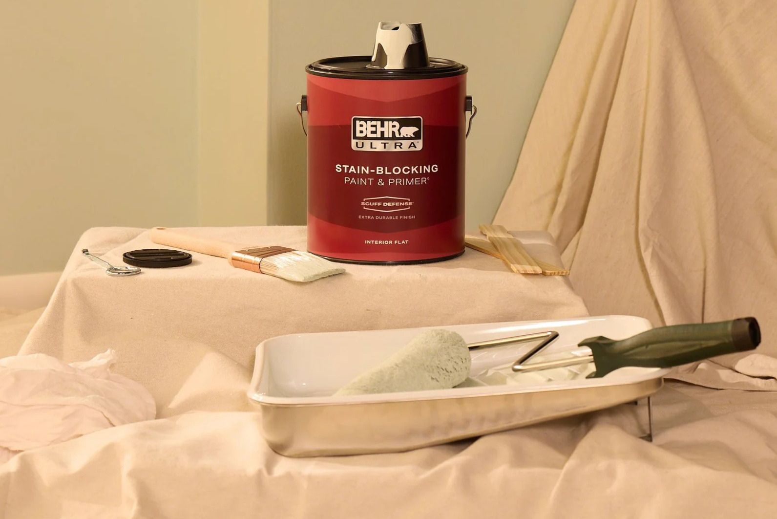 A can of stain-blocking paint sits on a covered surface alongside a paintbrush, a roller, and a paint tray, preparing for a painting project in a room with neutral walls, featured in the Behr $5,000 Sweepstakes