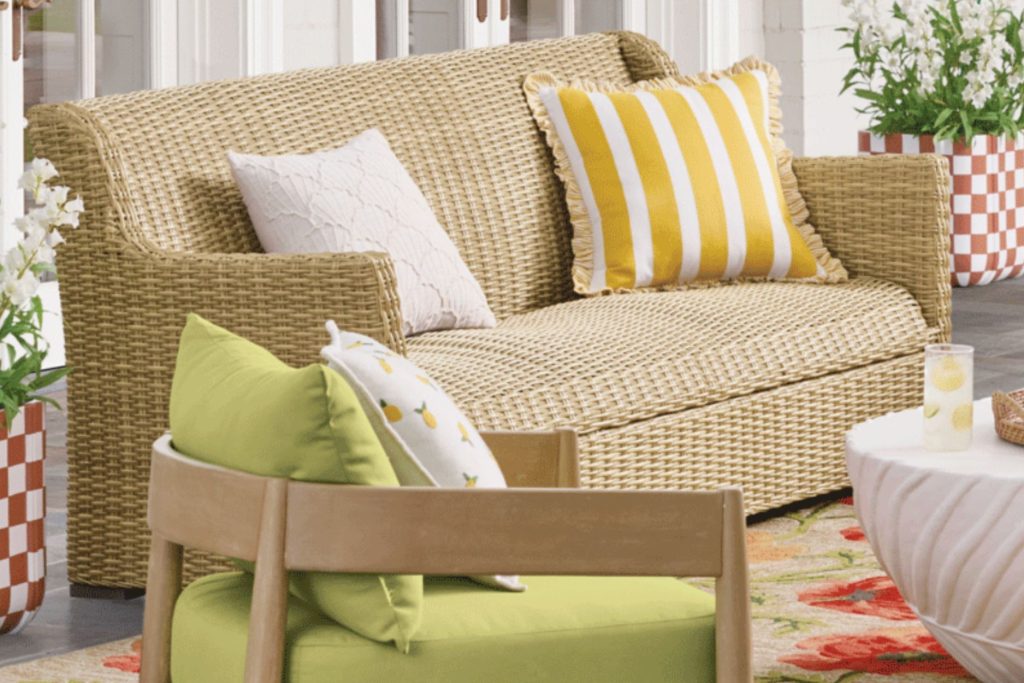 A stylish outdoor seating area featuring a woven beige sofa with decorative cushions, a green accent chair, and a round table with refreshing drinks on a floral rug, featured in the Grandin Road $5,000 Gift Card Sweepstakes