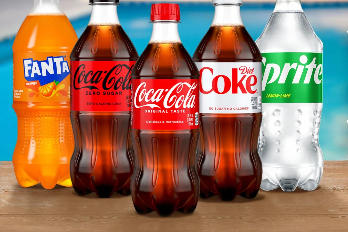 A selection of soft drink bottles sits on a wooden table, featuring orange soda, cola varieties, and a lemon-lime beverage, with a blue pool in the background, featured in the Coca-Cola Product Sweepstakes