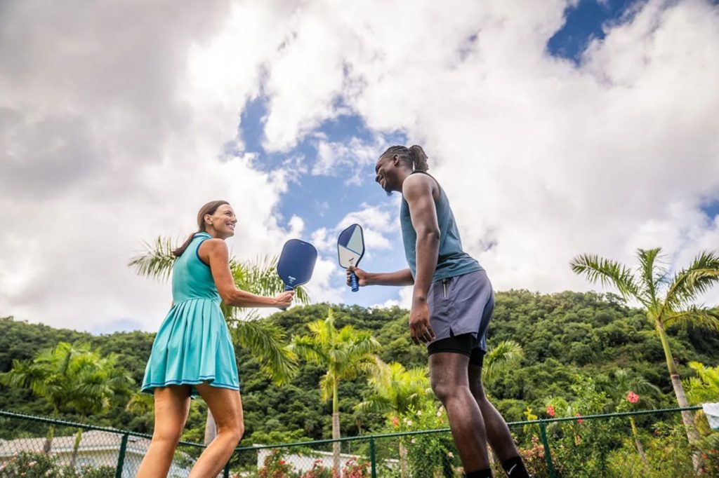 A young woman in a teal dress and a man in athletic wear playfully smile and hold pickleball paddles in a sunny outdoor setting, surrounded by palm trees and vibrant greenery, featured in the USA Pickleball Sandals Sweepstakes