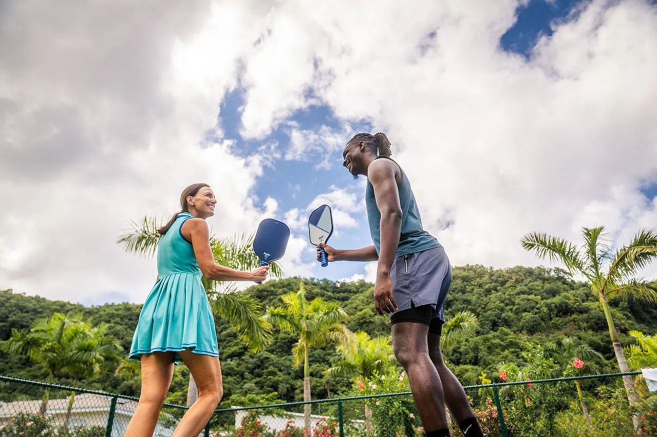 A young woman in a teal dress and a man in athletic wear playfully smile and hold pickleball paddles in a sunny outdoor setting, surrounded by palm trees and vibrant greenery, featured in the USA Pickleball Sandals Sweepstakes