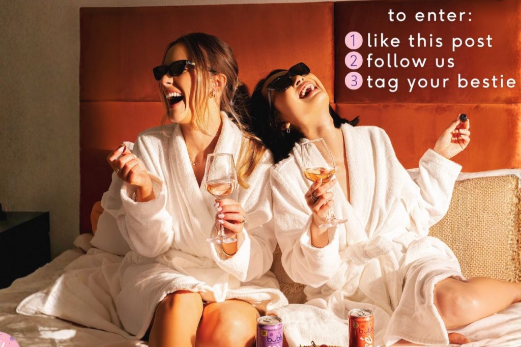 Two women in white bathrobes sit on a bed, laughing and enjoying wine glasses. One holds a snack, while colorful cans are visible nearby, capturing a joyful moment of friendship, featured in the Bubly Wine Refresher Trip Sweepstakes