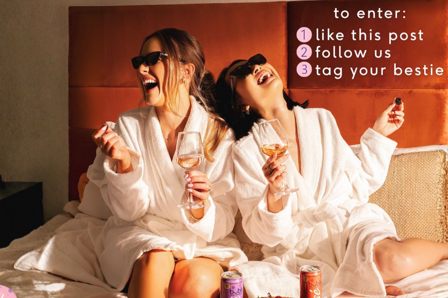 Two women in white bathrobes sit on a bed, laughing and enjoying wine glasses. One holds a snack, while colorful cans are visible nearby, capturing a joyful moment of friendship, featured in the Bubly Wine Refresher Trip Sweepstakes