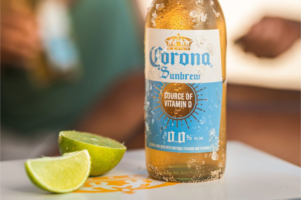 A chilled bottle of light beer sits beside freshly cut lime wedges on a table, capturing a refreshing summer vibe. The background hints at a relaxed outdoor setting, featured in the Corona Sunbrew Trip Sweepstakes