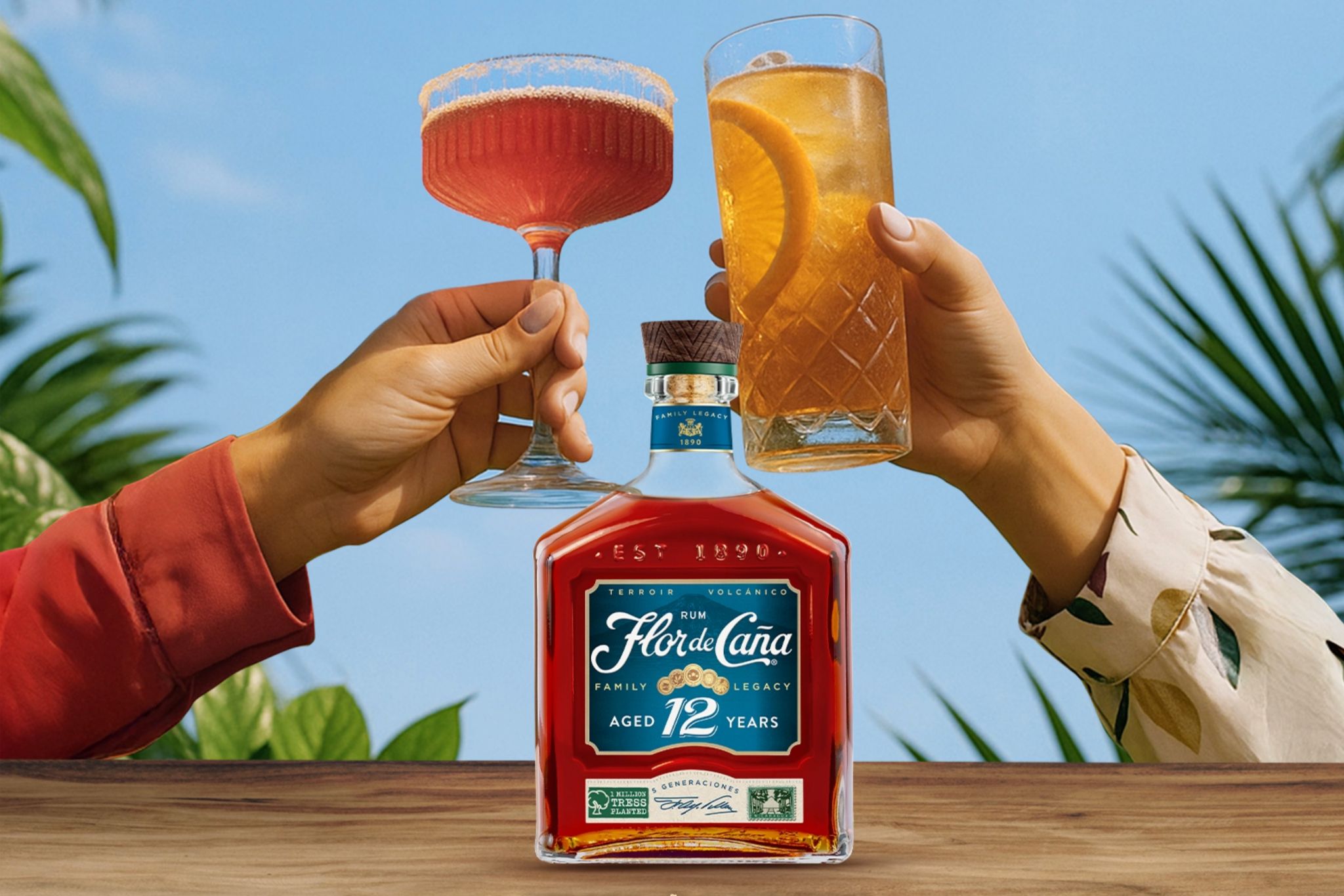 Two hands clink glasses in a tropical setting, one holding a pink cocktail and the other a golden drink with an orange slice, while a bottle of rum sits on a wooden table, featured in the Flor de Caña Costa Rica Sweepstakes