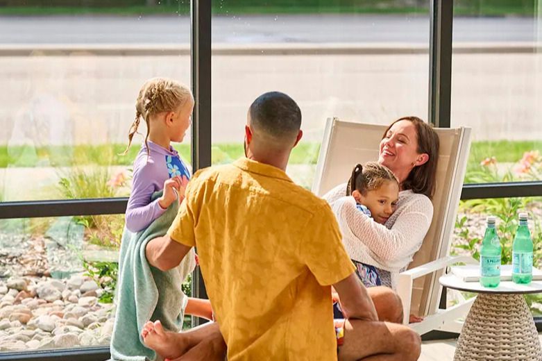 A joyful family enjoys a sunny day indoors. A woman cuddles a young girl while a man playfully interacts with another child, surrounded by greenery and light, featured in the Bloomington Mother's Day Sweepstakes