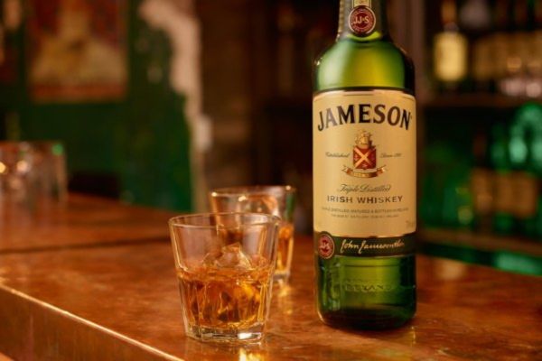 A bottle of Irish whiskey stands on a polished wooden bar top next to two glasses filled with whiskey and ice, surrounded by a warm, inviting setting, featured in the Jameson Goal-Den Ticket Sweepstakes