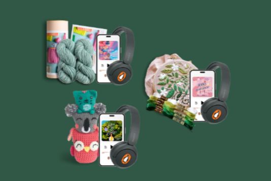A collection of colorful yarn, knitting supplies, and crocheted items arranged with phones and headphones, showcasing a cozy crafting theme on a green background, featured in the Maker’s Magic March 2026 Audio Crafting Giveaway